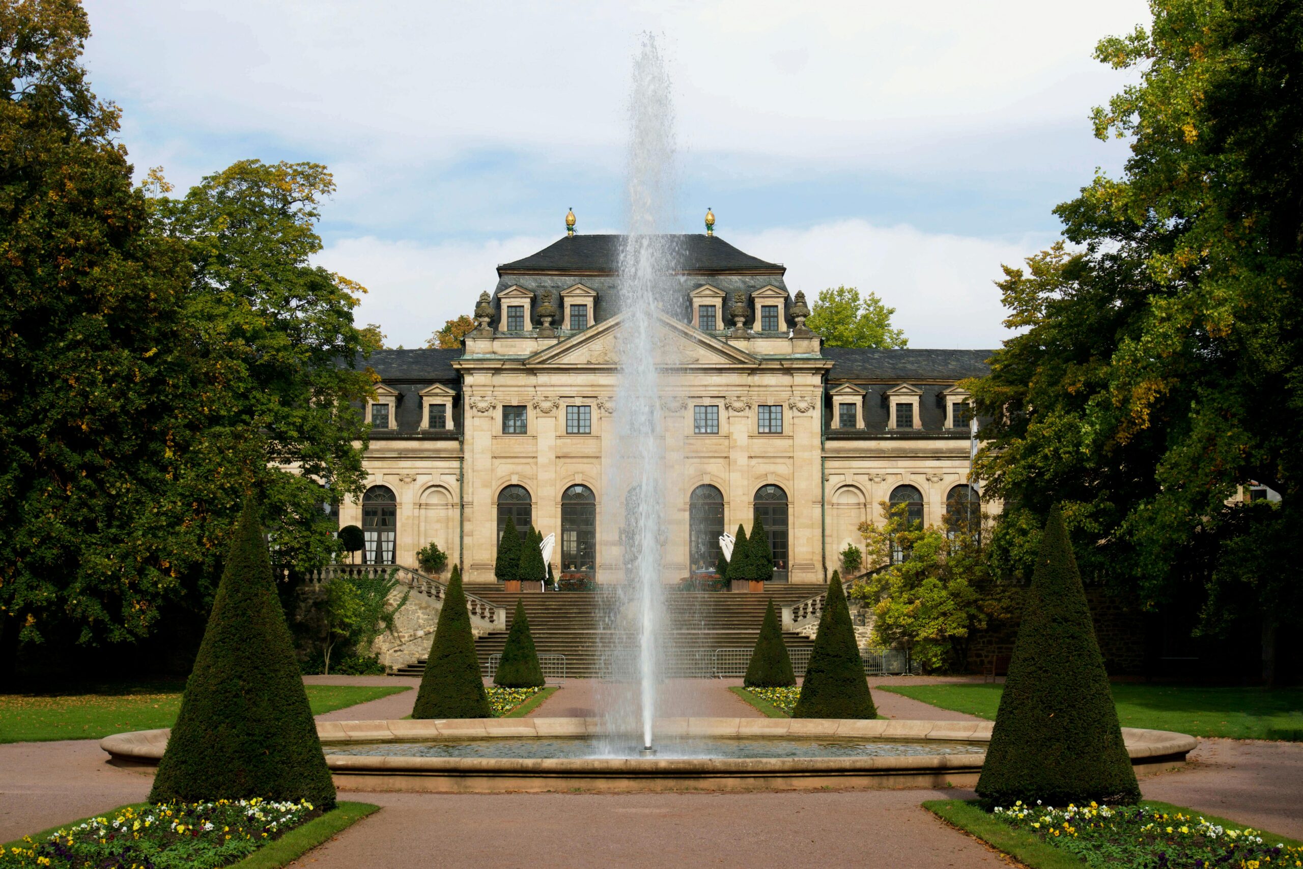 Elegant historic mansion facade with central fountain surrounded by lush gardens, offering a glimpse of classic architecture.